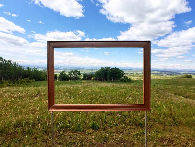 View from the Leighton Art Centre, with the Rocky Mountains in the distance and grassy hills with trees. An empty frame is suspended in the middle, showing the view beyond as if in a painting.