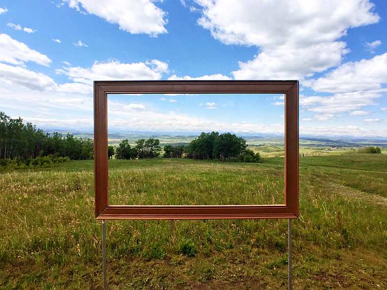 View from the Leighton Art Centre, with the Rocky Mountains in the distance and grassy hills with trees. An empty frame is suspended in the middle, showing the view beyond as if in a painting.